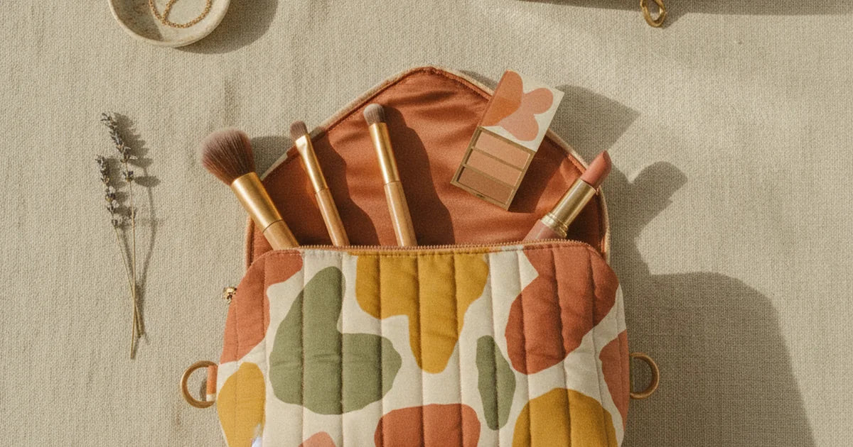 hero — trousse de maquillage matelassée à imprimé coloré posée ouverte sur une coiffeuse claire, pinceaux, rouge à lèvres et palette qui dépassent, miroir et lumière douce du matin, ambiance beauté soignée