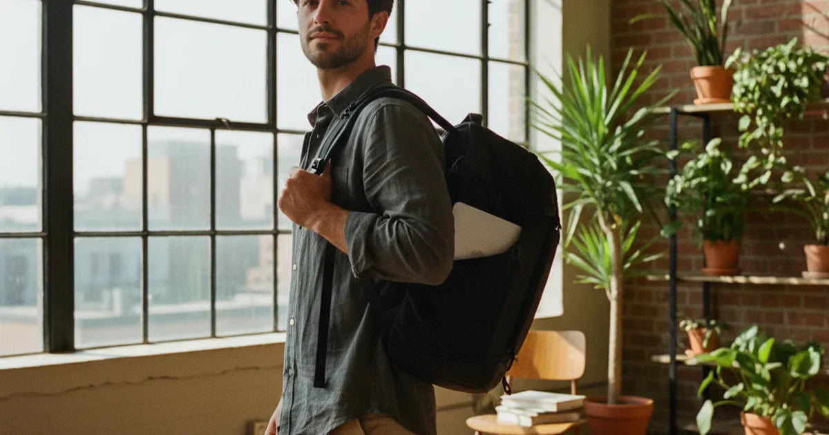 sac à dos voyage — jeune femme en chemise oxford crème portant un sac à dos technique 40 litres noir avec ordinateur portable accessible, à un quai de gare ensoleillé avec un train à l'arrière-plan