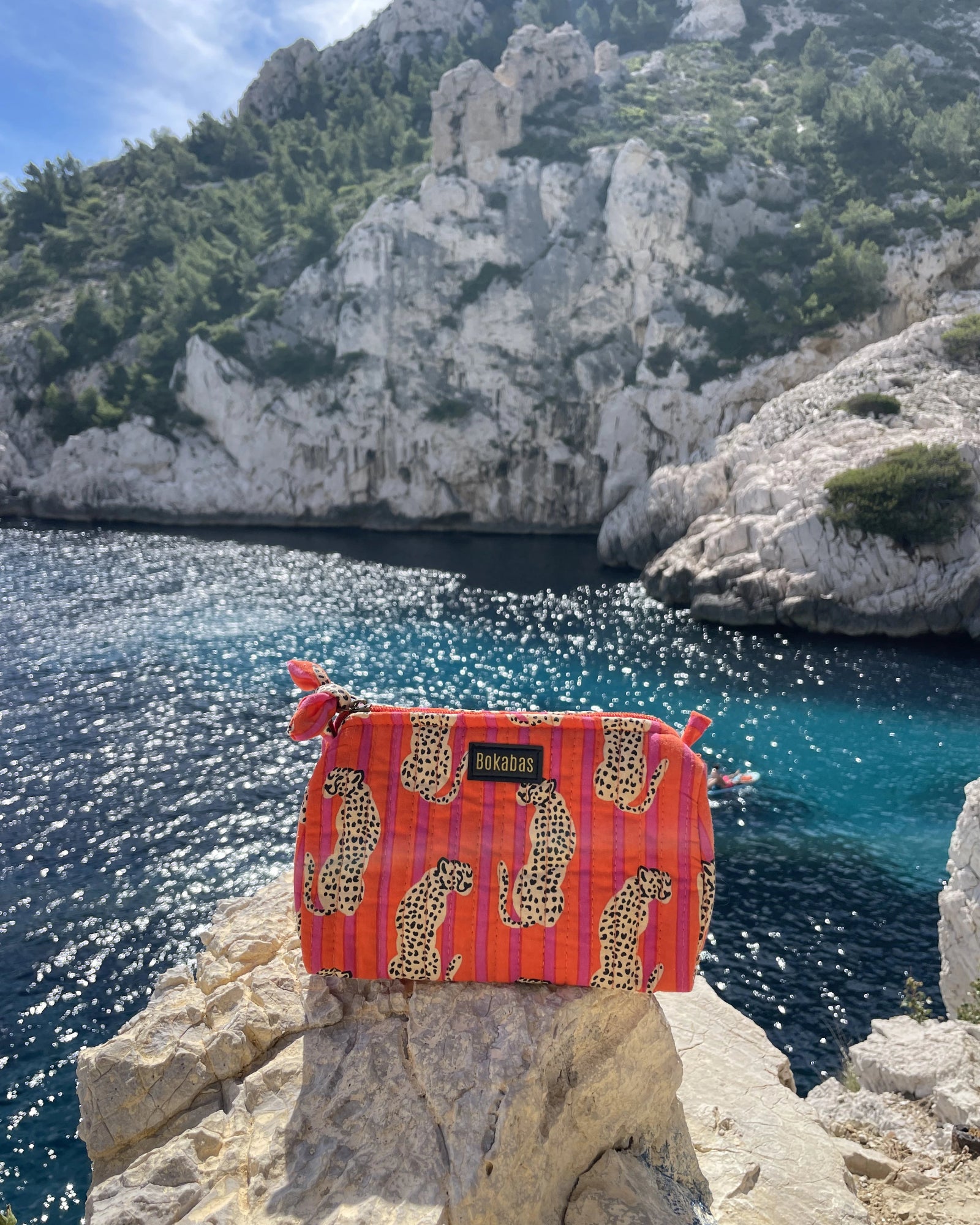 Trousse de toilette Bokabas, imprimé Guépard orange et rose, posée sur un rocher face à la mer